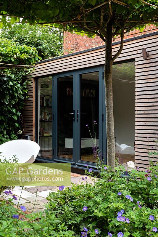 View across patio to modern summerhouse from under trained tree arch. 