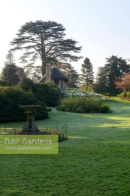 View across lawn with island beds towards thatched Swiss Cottage with trees beyond