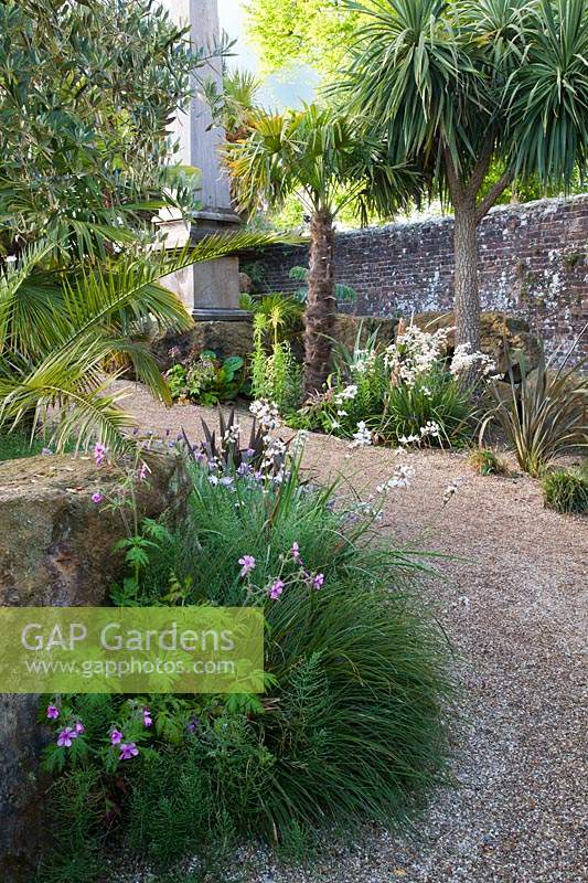 Path towards stone monument, with structural palms, grasses, Libertia, Geranium and olive trees planted around rockery. Arundel Castle, West Sussex, UK. 