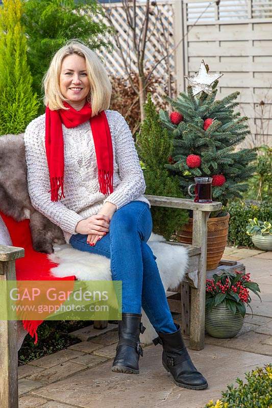 Woman sat on bench with sheepskins having a warm drink, surrounded by Christmas greenery and decorations