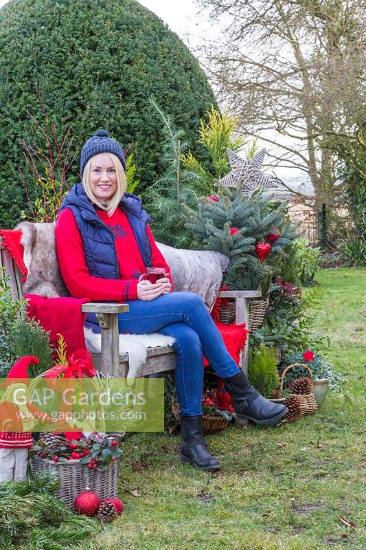 Woman sat on bench with sheepskins having a warm drink, surrounded by Christmas greenery and decorations