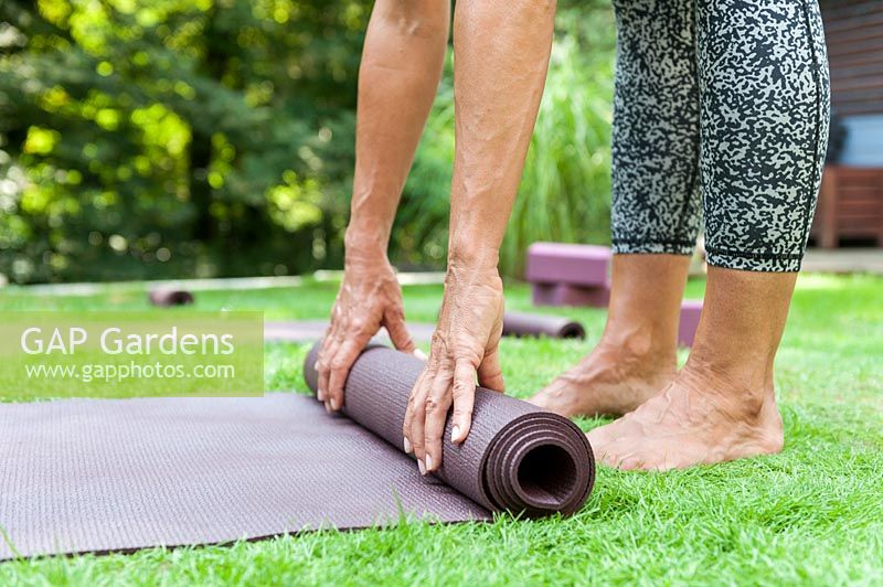 Woman standing barefoot in garden, rolling up yoga mat. 

