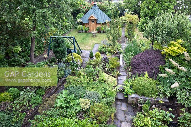 Overview of enclosed city garden from the house showing full planting beds, paved paths and Swedish summerhouse or sauna
