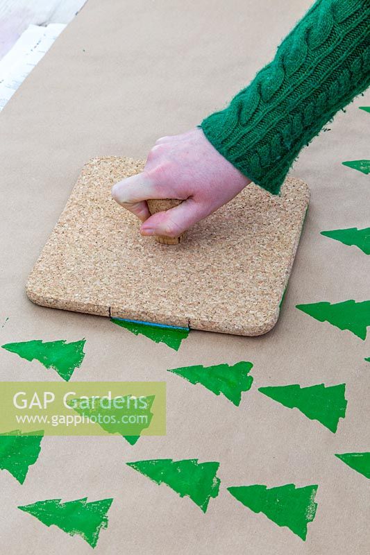 Woman creating continuous christmas tree pattern onto brown wrapping paper with cork stamp.