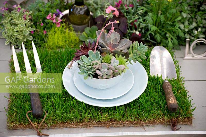 Garden table setting, with grass table mat, garden fork and trowel and succulent filled bowl. 
