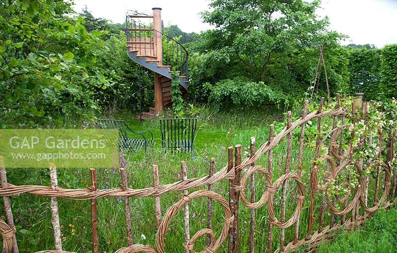 Hazel and willow decorative fence in pattern of circles with wildflower meadow beyond, and spiral staircase in the show garden 'Belmond Enchanted Gardens' 