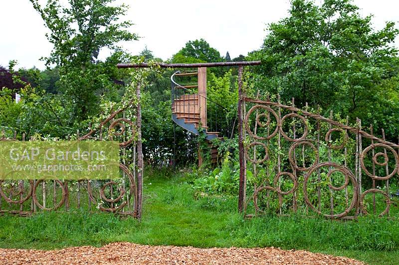 Hazel and willow decorative fence in pattern of circles, through a gap a spiral staircase in the show garden 'Belmond Enchanted Gardens'