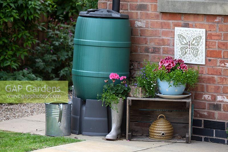 Water butt on stand by side of house with watering can, decorative crate, container plantings and wall plaque

 and planters
