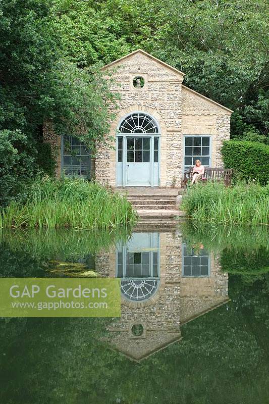 View across water and marginal planting to The Shell House Folly, reflected in the water
