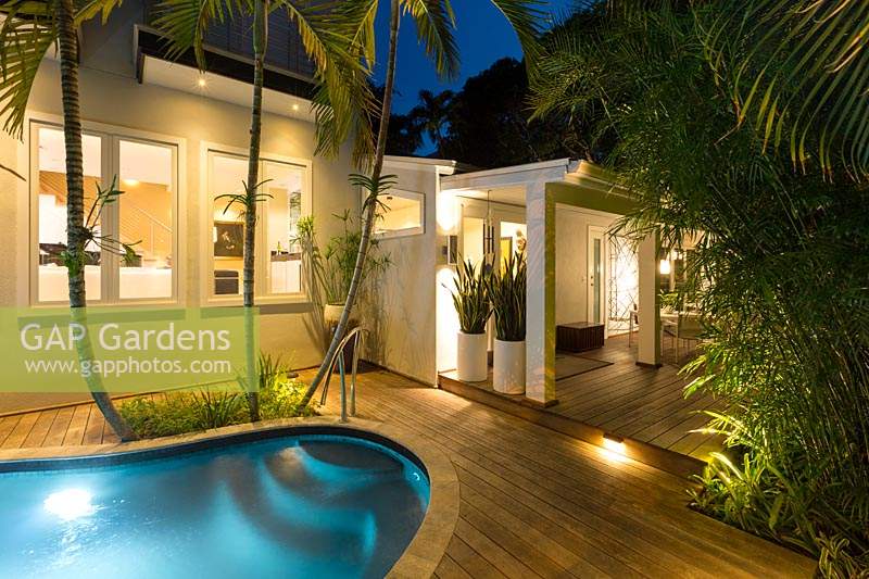 Tropical garden and house lit up at night, view from swimming pool and wooden deck towards entrance with pots of Sanseveria