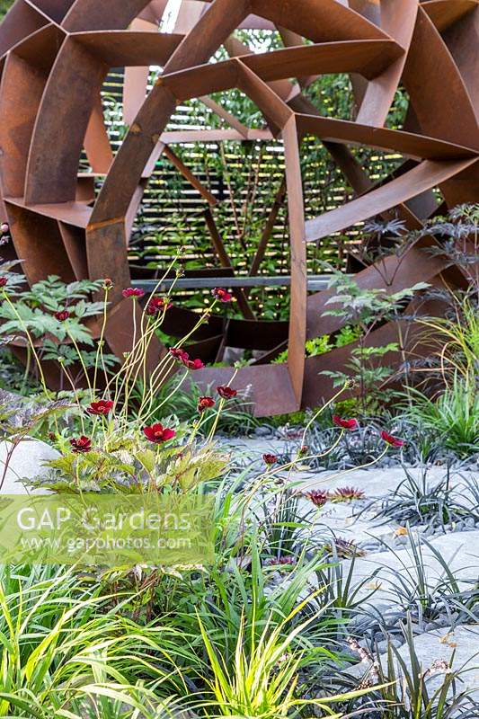 Corten steel sphere by William Roobrouck near ornamental grasses set in paving 
in 'Elements Mystique Garden', Sponsored by Elements Garden Design