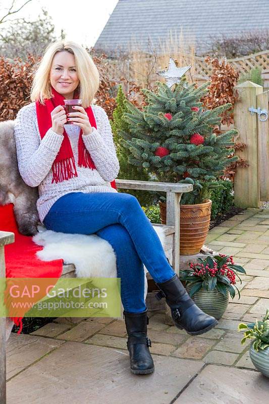 Woman relaxing on bench with sheepskins, a decorated Christmas tree in background having a hot drink. 