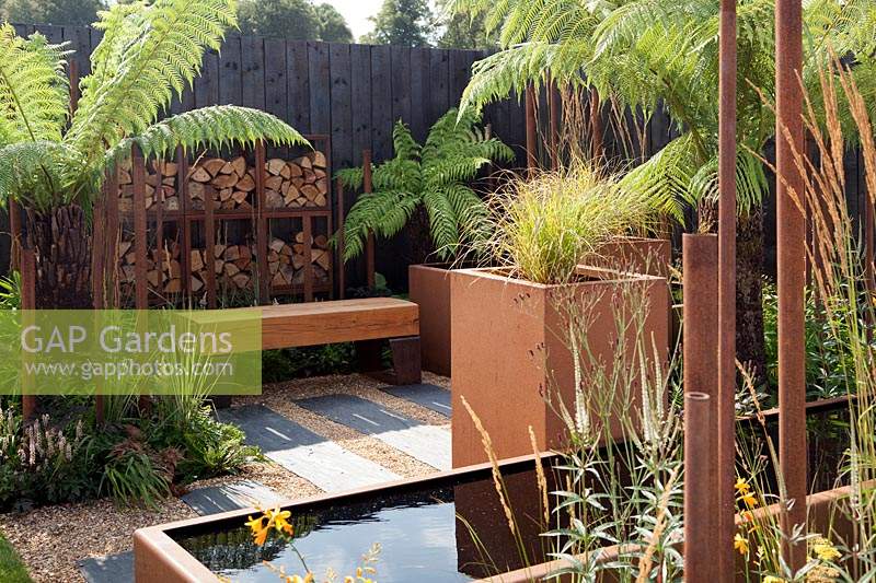 Dicksonia antarctica in contemporary, urban style garden with Corten water trough and planters and chunky oak bench in 'The Penumbra' garden, RHS Tatton Park Flower Show 2018