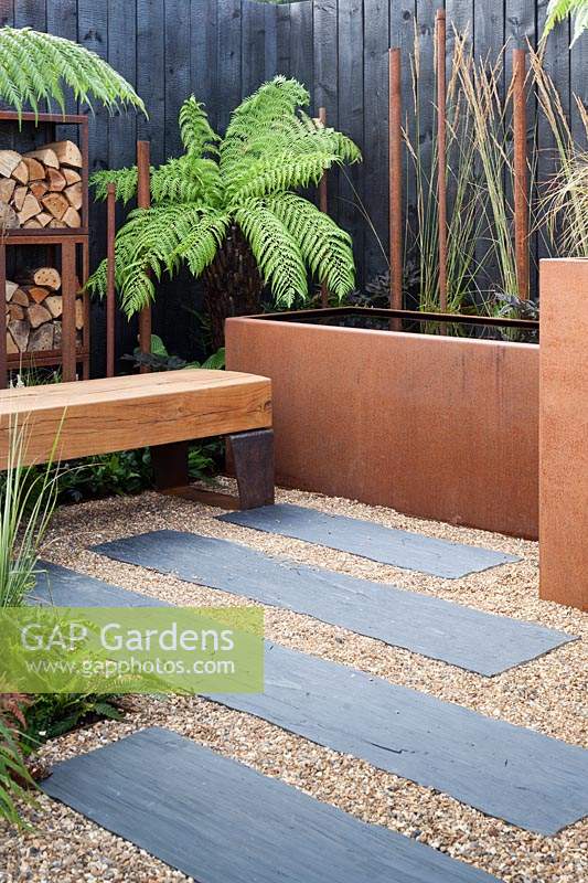 A chunky oak bench, Corten water trough and log storage in a contemporary, urban style garden in 'The Penumbra' garden, RHS Tatton Park Flower Show 2018