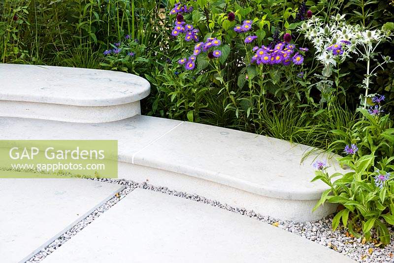 Marble bench surrounded by mixed borders of Erigeron 'Darkest Of All', Centaurea montana, Allium sphaerocephalon and Eryngium giganteum. The South West Water Green Garden at RHS Hampton Court Palace Flower Show 2018 