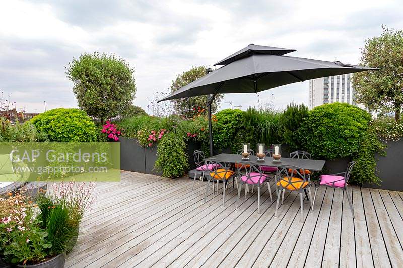 London roof terrace with wood decking and contemporary grey chairs, table and parasol. In the background raised beds with perennials and annuals forming a natural screen for privacy.
