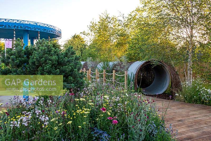 Wooden path leading to a tunnel made from concrete drainage pipes, perennial meadow with Penstemon 'Husker Red', Lychnis coronaria, Echinacea, Achillea and Verbena bonariensis next to Pinus mugo. RHS Hampton Court Palace Flower Festival 2019.
