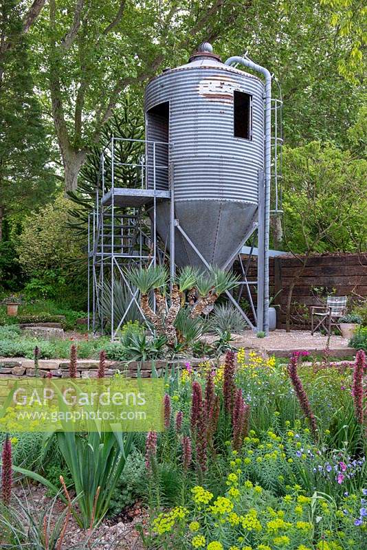 Repurposed grain silo used as an office, planting in foreground of Digiplexus, Echium russicum, Euphorbia palustris with Yucca recurva behind - The Resilience Garden, RHS Chelsea Flower Show 2019.