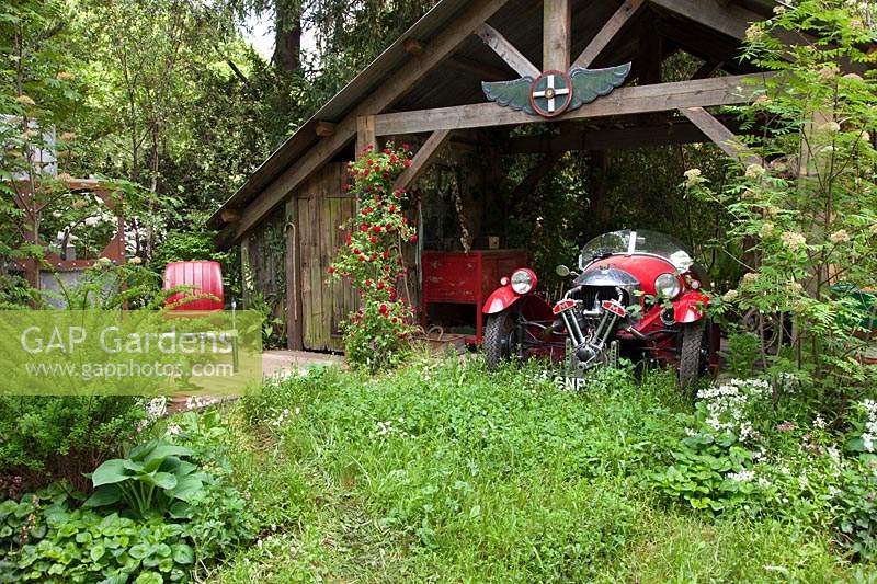 The High Maintenance Garden for Motor Neurone Disease Association, view of the garden where a garage with a hand-built sports car is being reclaimed by nature, the planting includes red rambling rose, Hosta and Primula. Sponsor: MND Association.