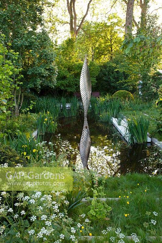 The Savills and David Harber Garden - View of the pool with the sculpture representing a leaf and surrounded with a woodland clearing, the planting includes Anthriscus sylvestris, Tellima grandiflora, Iris pseudacorus, and Ranunculus acris - Sponsor: Savills and David Harber.