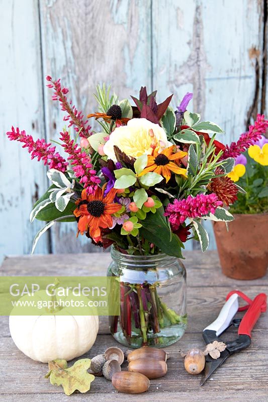 Posy or Tussie Mussie with a colourful mix of flowers and foliage in a jam jar. On a wooden table with nuts, Winter Squash and secateurs