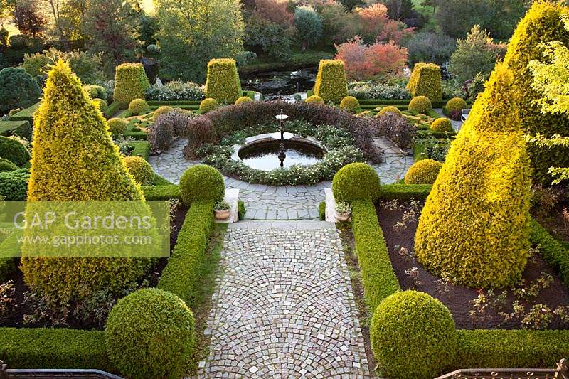 View from the house onto the Terrace with fountain. Four cut off pyramids of Thuja occidentalis Sunkist in background and two cones of same in foreground. Low hedge of Spirea japonica Little Princess by path. Orekhovno garden, Orekhovno, Pskov Oblast region, Western Russia. 