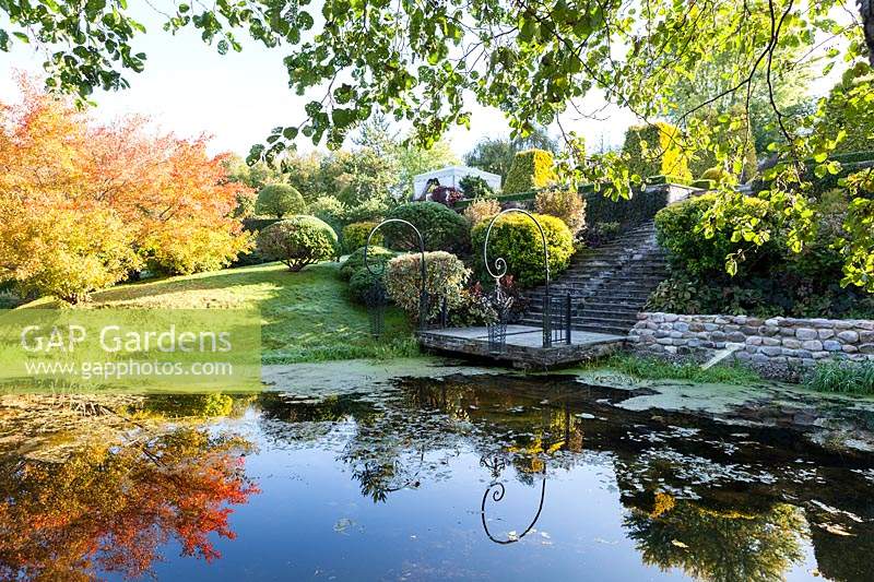 The landing with ornate hanging lanterns viewed from the Orekhovnitza river and reflected in the water. Acer ginnala with autumn colour on left. Orekhovno garden, Orekhovno, Pskov Oblast region, Western Russia.