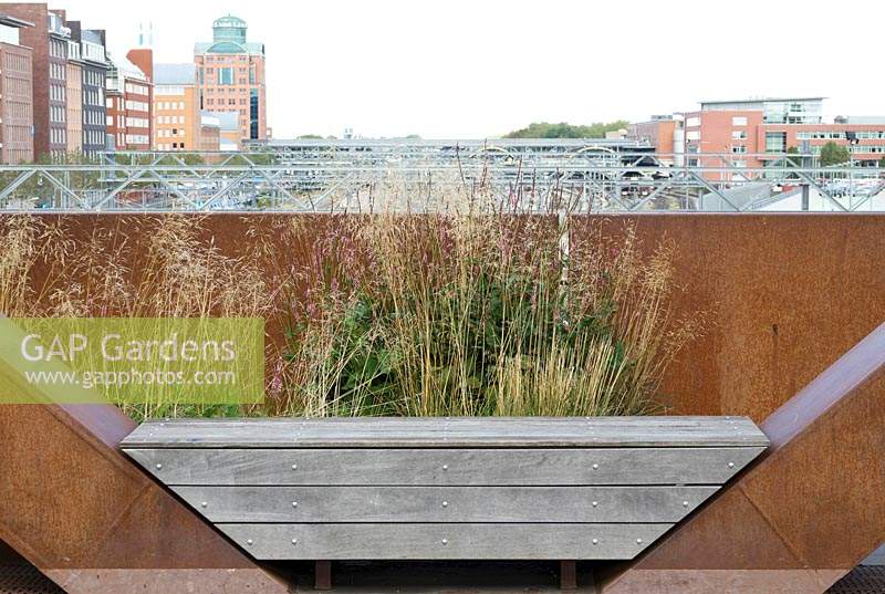 Wooden bench between corten steel wall with view of the railway and city buildings.