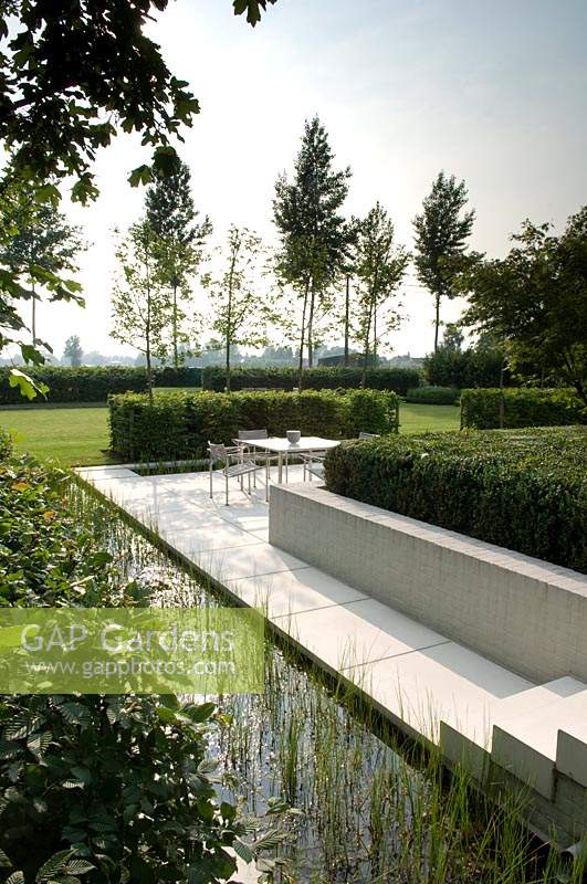 Long rill alongside paved patio with dining area in modern garden. 