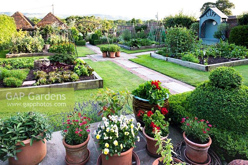 View of kitchen garden from terrace edged with container plants. Kitchen garden divided into quarters by paved paths, each quarter has lawn with large raised beds edged with railway sleepers, blue arbour along boundary fence