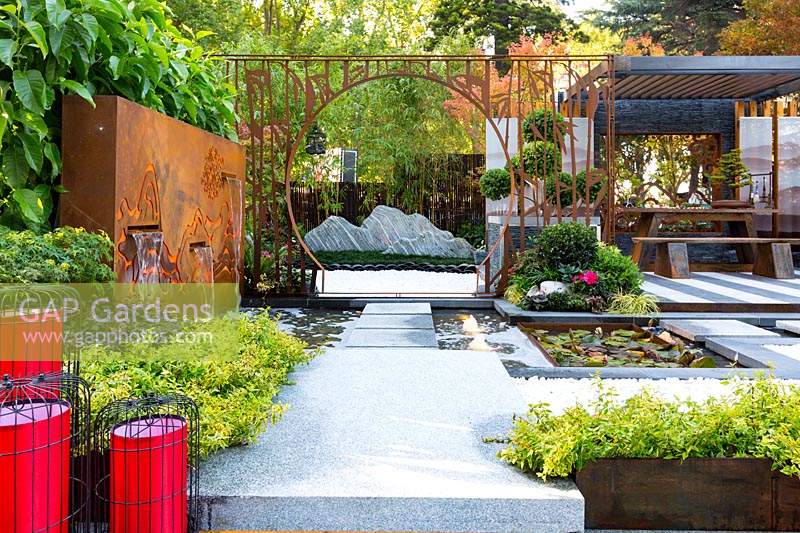 Pavilion style Chinese garden, with a freestanding water feature, moongate, stepping stones, striped paving , planter boxes, stone mountain range, printed glass panels, steps and red lanterns.