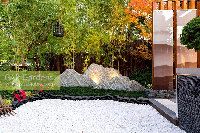 View of rocks representing a mountain range, across stepping stones. A green screen of golden bamboo and a dark brown bamboo fence.