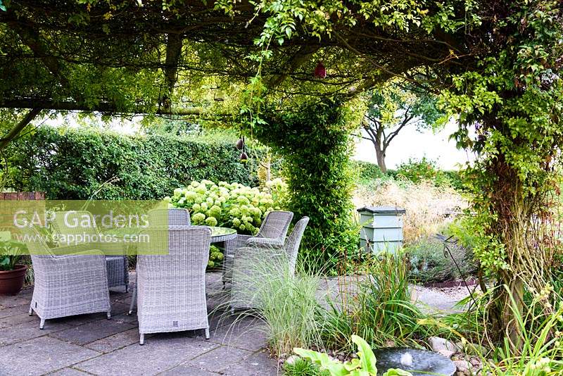 Rose covered arbour over a dining area in a rural garden with bubble millstone water feature in the foreground and grasses and hydrangea behind.