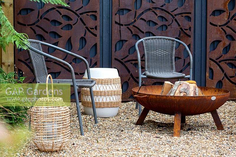 With gravel patio, and chairs around a fire pit. In background decorative rusty metal panels on wooden fence.