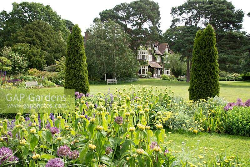 A view of Abbeywood House, an Edwardian residence built in 1908, seen accross a herbaceous border with  Allium cristophii Achillea 'Moonshine'and Phlomis russeliana and repeated conifers.