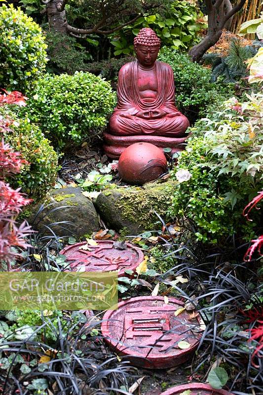 Red buddha statue set amongst beds autumnal colours and stepping stones at Four Seasons garden, Walsall, West Midlands.