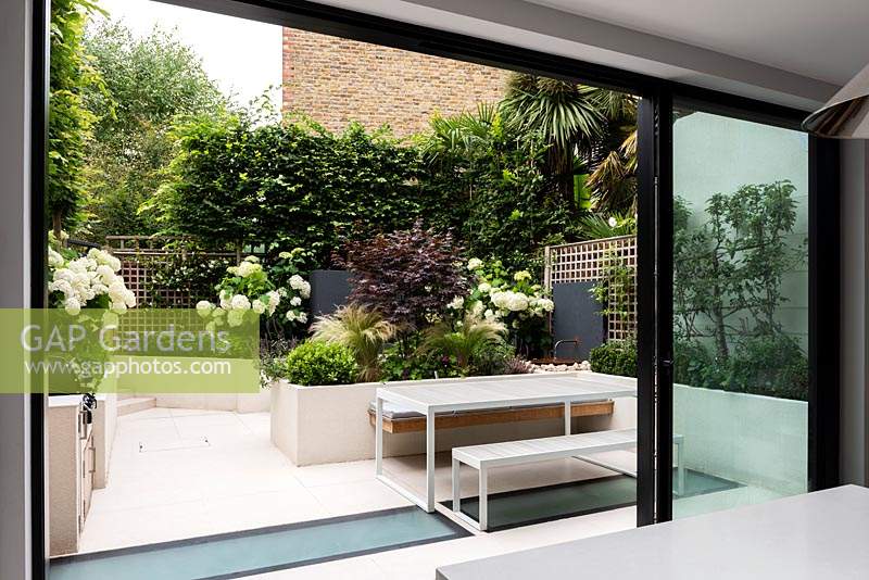 View from the kitchen through sliding glass doors to an outdoor dining area and raised beds. Bordering Carpinus Betulus gives the space privacy.