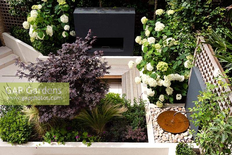 View from above of small contemporary private urban garden with porcelain paving, raised beds with planting of Hydrangea arborescens 'Annabelle', Acer palmatum 'Bloodgood' and Stipa tenuissima, outdoor fireplace, floating bench seating and a copper pipe and bowl water feature.