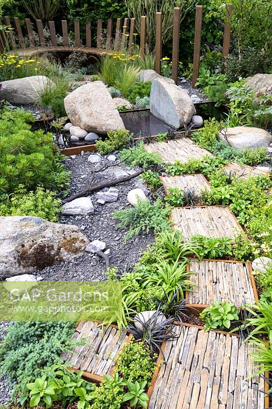 Natural rock and stone, with green foliage groundcover planting. Wooden seating and  stepping stones with slate pieces in corten steel frames with Deschampsia cespitosa, Achillea 'Moonshine', Ophiopogon planiscapus 'Nigrescens' - Through Your Eyes Garden - RHS Hampton Court  Palace Garden Festival 2019. Sponsors: Kebony, CED Stone, R and G Metal Products, William's Art and Design, Practicality Brown.