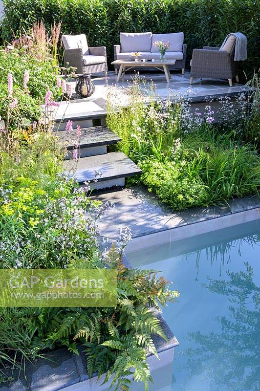 Stone steps from pool leading to raised patio with outdoor sofa and armchairs - APL - A Place To Meet Garden - Hampton Court Palace Garden Festival 2019.