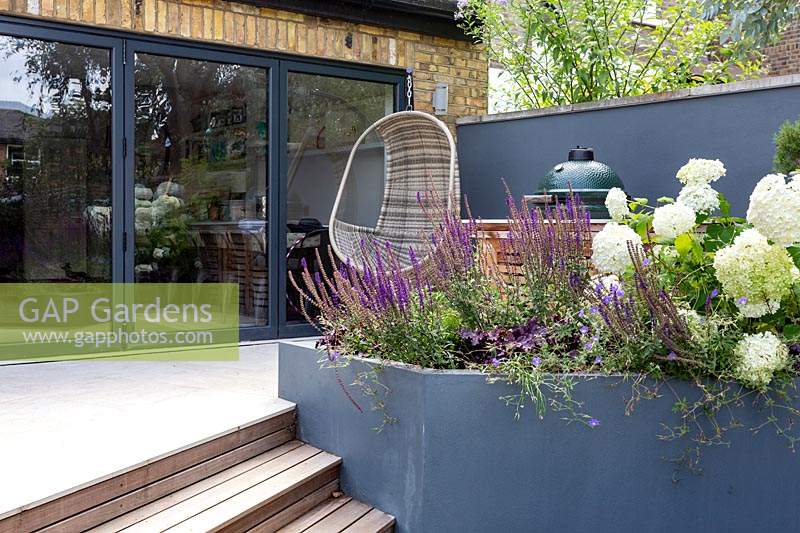 London contemporary garden with view towards house - bespoke barbecue on patio with grey raised border. Planting includes Heuchera berry smoothie, Salvia caradonna, Hydrangea anabelle, Geranium johnsons blue.