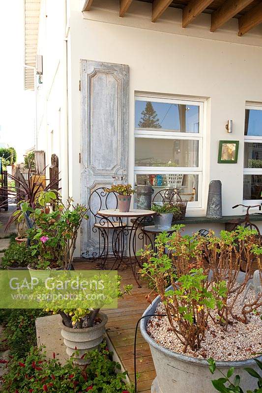 Corner of a verandah with rusty metal outdoor table and chairs and potted, mismatched a retro metal bucket with a succulent and a metal tub with a Pelagonium in it in front of a shabby chic panelled door.