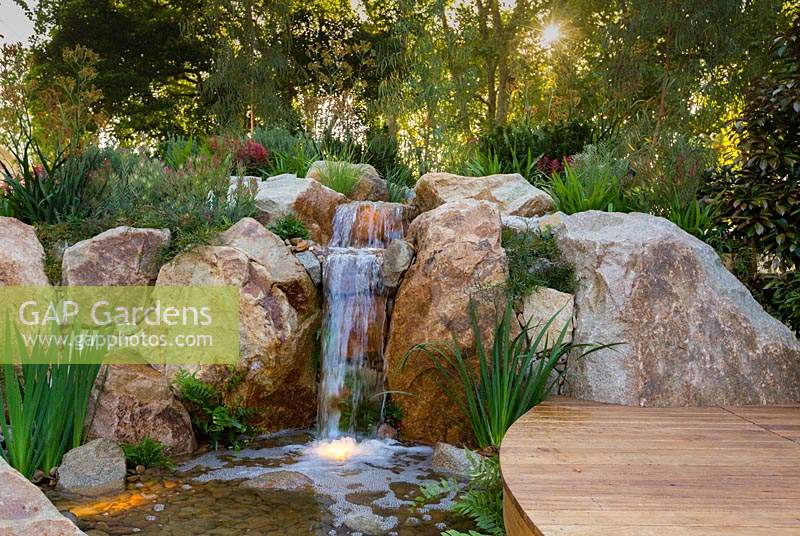 A waterfall constructed from large boulders and small rocks, planted out with grasses, flowering plants and groundcovers and featuring a curved hardwood timber boardwalk