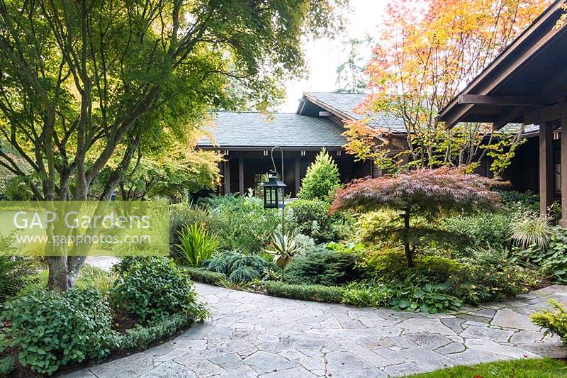 A flagstone path leads to the Craftsman-style home, stained dark brown to blend into its surroundings. Garden beds adjacent to the home are filled with an eclectic collection of trees, conifers, shrubs, perennials and ornamental grasses 