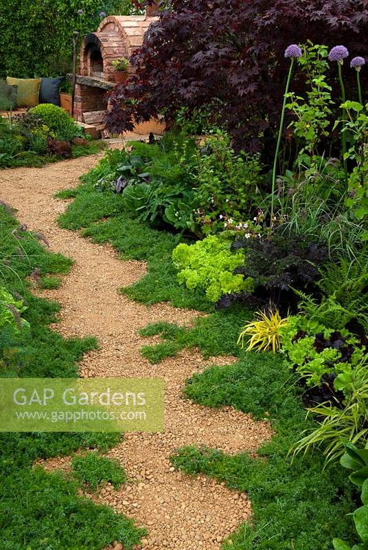 Chamomile edged gravel path through mixed borders of vegetables, herbs, ferns and salad crops leading to sunken area with outside oven and seating - RHS Malvern Spring Festival 2014