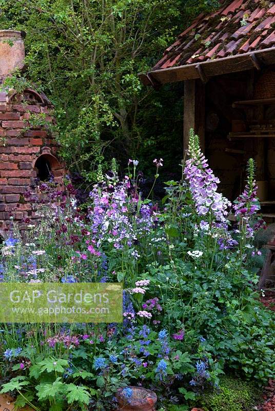 Herbaceous plants and wildflowers in cottage garden style border with rustic brick kiln and workshop beyond - RHS Chelsea Flower Show
