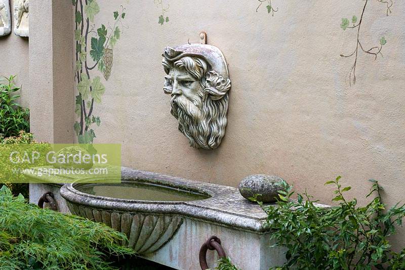 A horse trough wall fountain with Nepture's head above in a shady garden corner. 