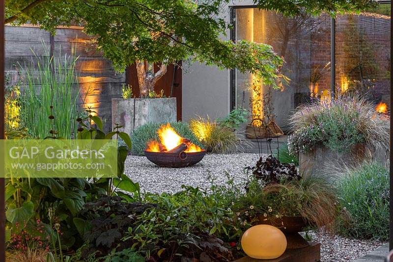 Seen through a window of foliage, a flaming fire pit reflected in the house windows.