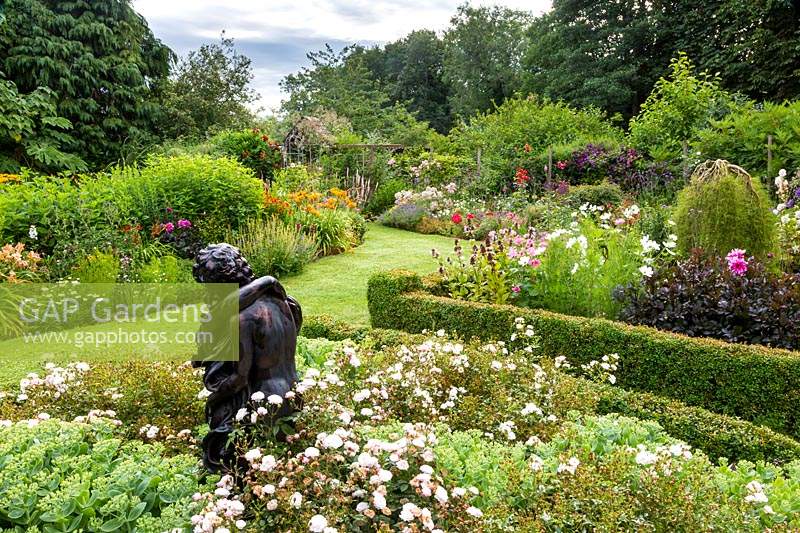 Packed flower beds either side of grass path, a statue of entwined lovers is among Rosa 'Little White Pet', Rosa 'Kent' and Sedum spectabile in the foreground