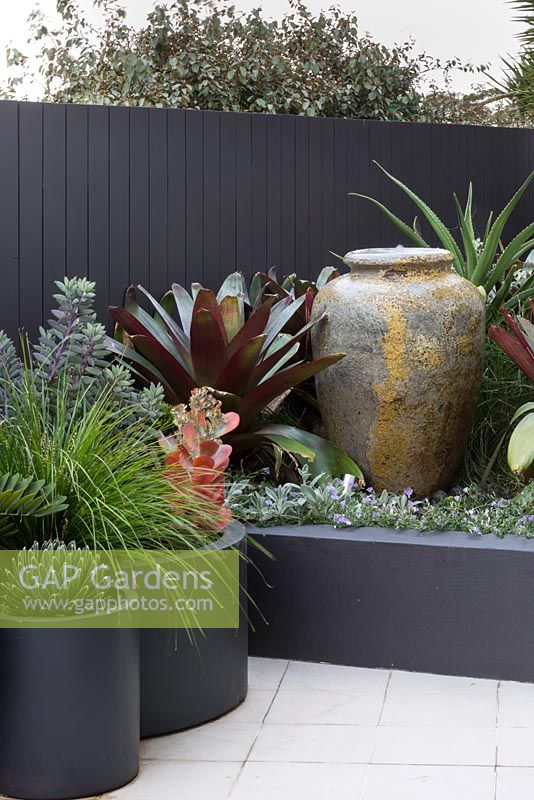 A large amphora shaped water bowl in a raised garden bed with a large bromeliad next to a group of cylindrical pots planted with a variety of plants.